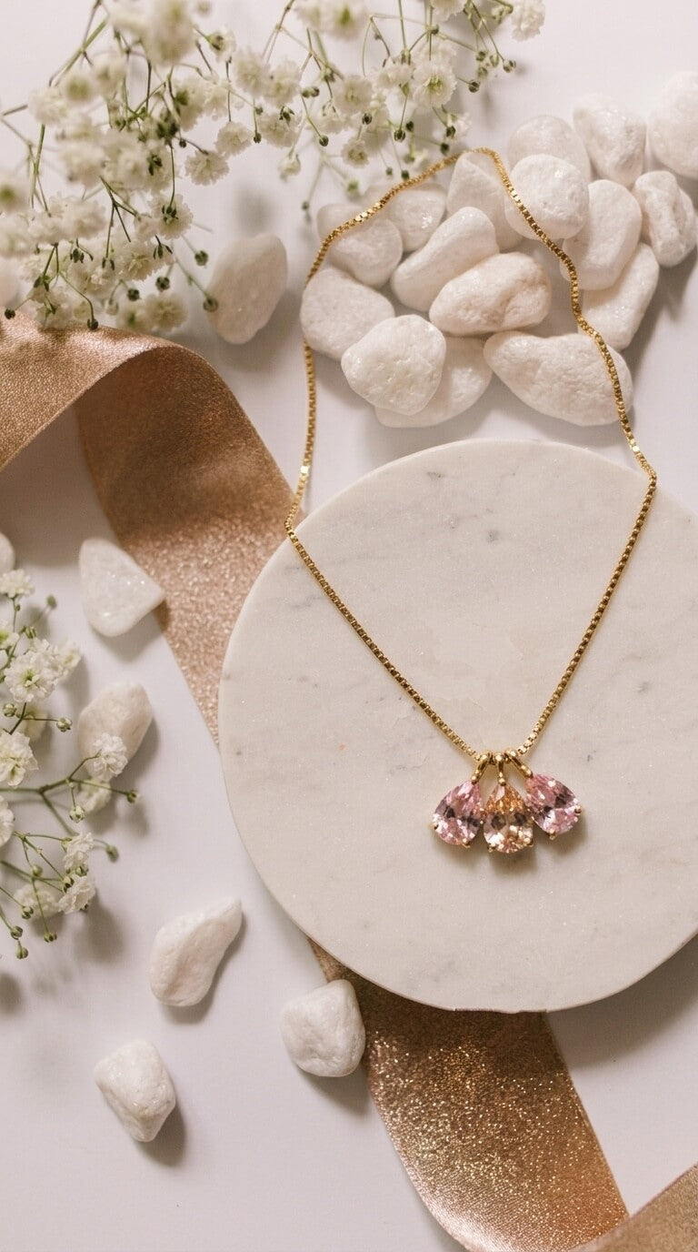 A gold chain necklace with a multi-stone pendant featuring pink, peach, and yellow hues, displayed on a round white marble coaster surrounded by white stones, ribbon, and baby's breath flowers.