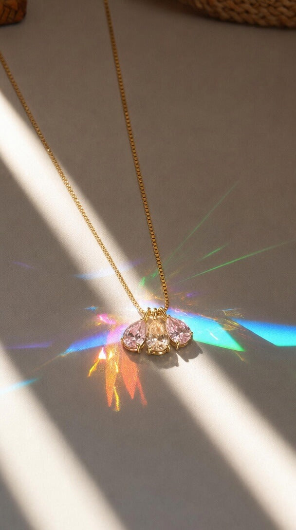 A delicate gold chain necklace with a butterfly-shaped crystal pendant, resting on a neutral surface with dramatic sunlight and colorful prism reflections.
