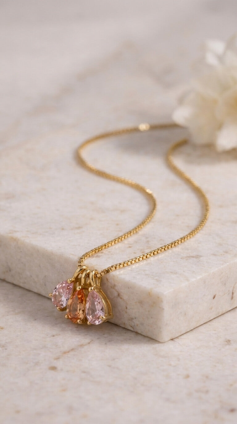 A delicate gold chain necklace with a cluster of teardrop-shaped pink gemstones, displayed on a light beige marble block with a soft white flower in the background.